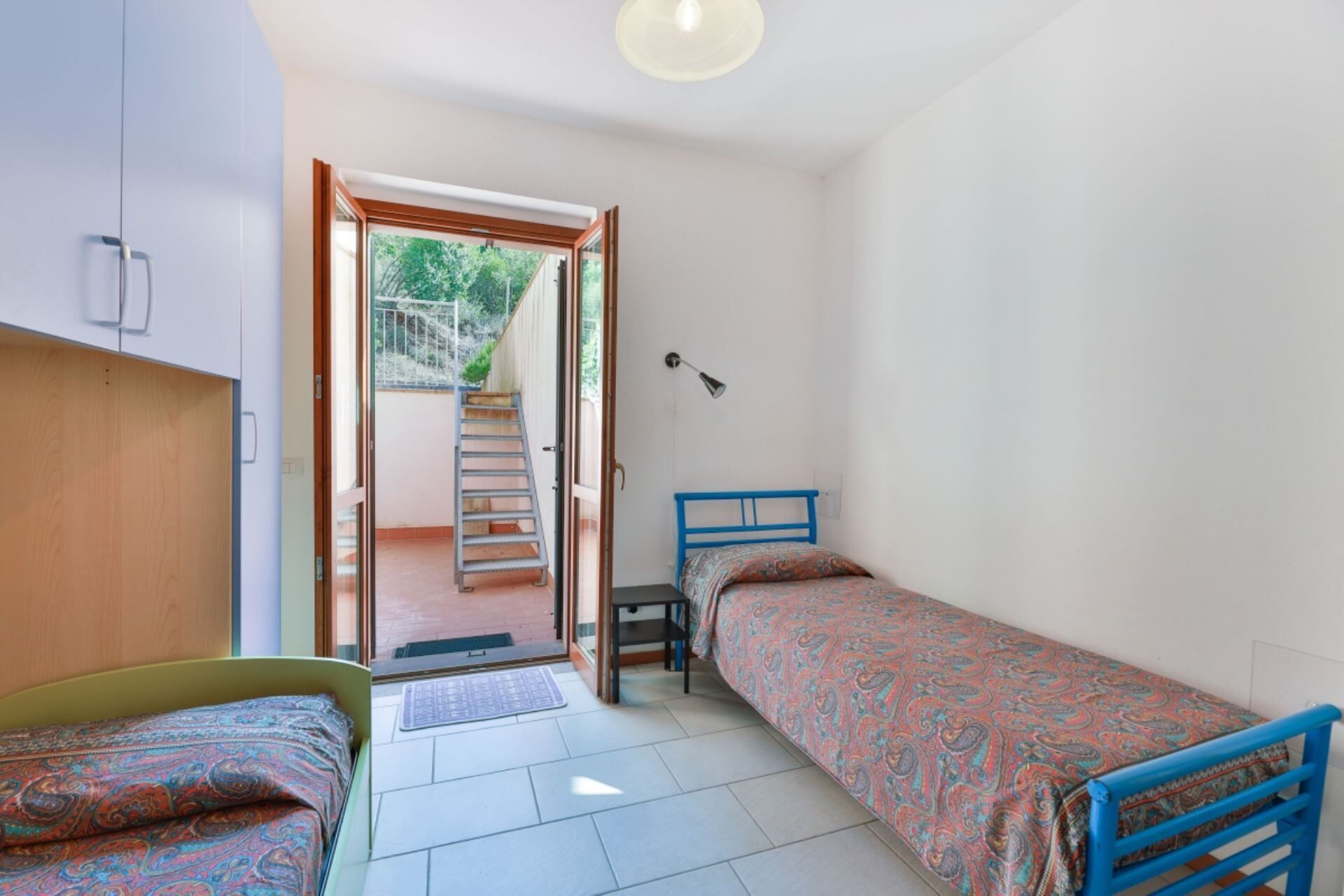Ein kleiner, gut beleuchteter Raum mit einem Bett, einem Stuhl und einem Schreibtisch. Eine Tür führt zu einer Treppe und es gibt einen Blick auf Grünflächen.
