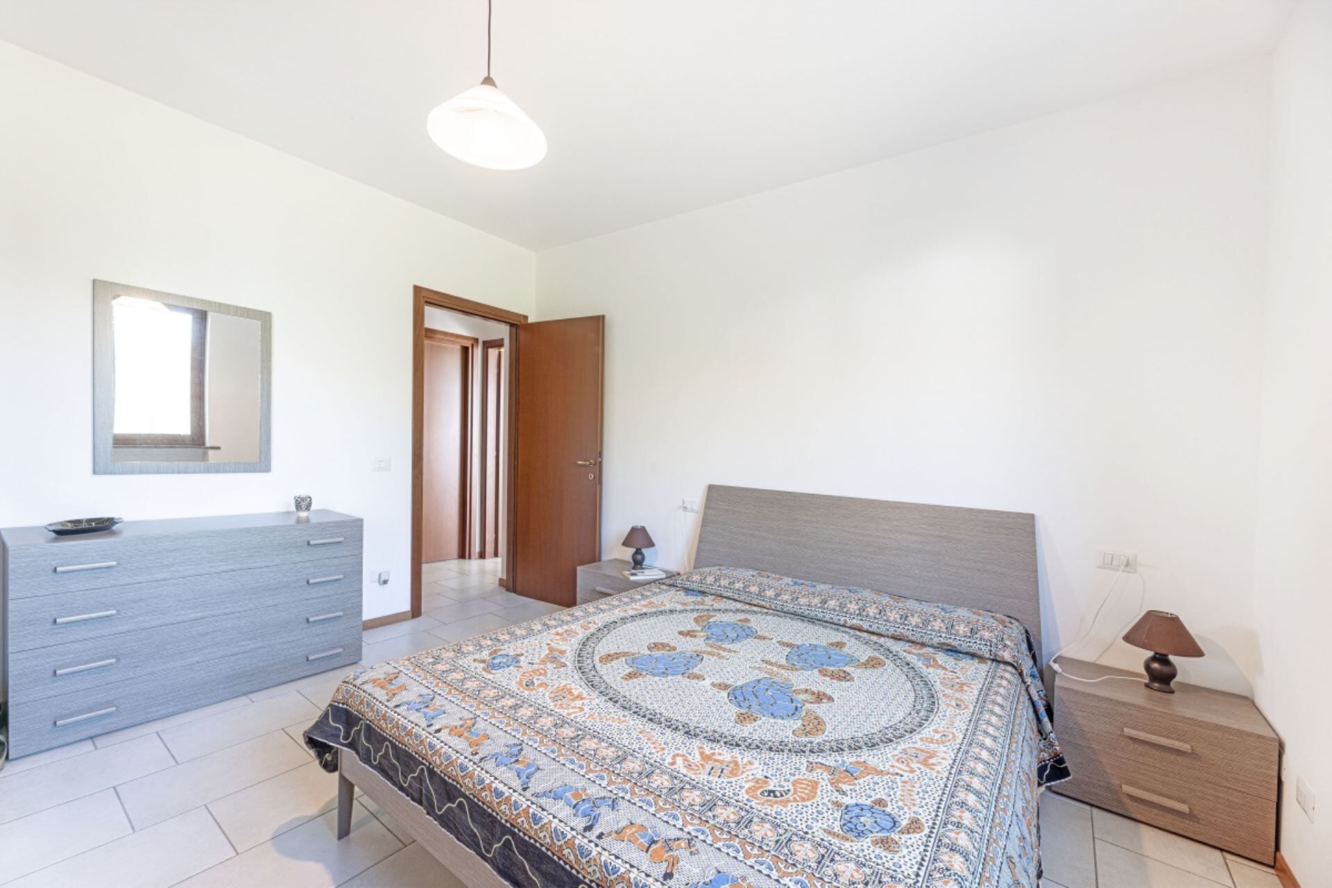 A bedroom with a bed, bedside tables, a dresser, a mirror, and a door leading to another room.