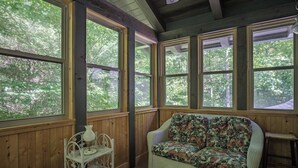A room with a wicker sofa, a small table with a lamp, and large windows with a view of trees. - Black Bear Lodge Near Dupont State Forest (Hendersonville)