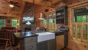 A rustic cabin interior with a stone fireplace, wooden ceiling, and a kitchen area with a sink and cabinets. - Black Bear Lodge Near Dupont State Forest (Hendersonville)