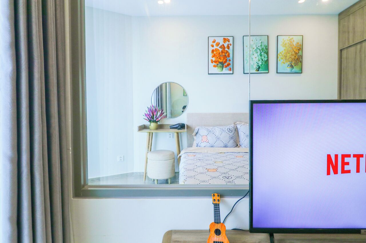 A modern bedroom with a large window, a bed, a TV displaying 'NETFLIX', a guitar, and framed floral artwork on the wall.