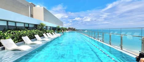 A long, clear swimming pool with white lounge chairs on one side and a glass railing on the other.