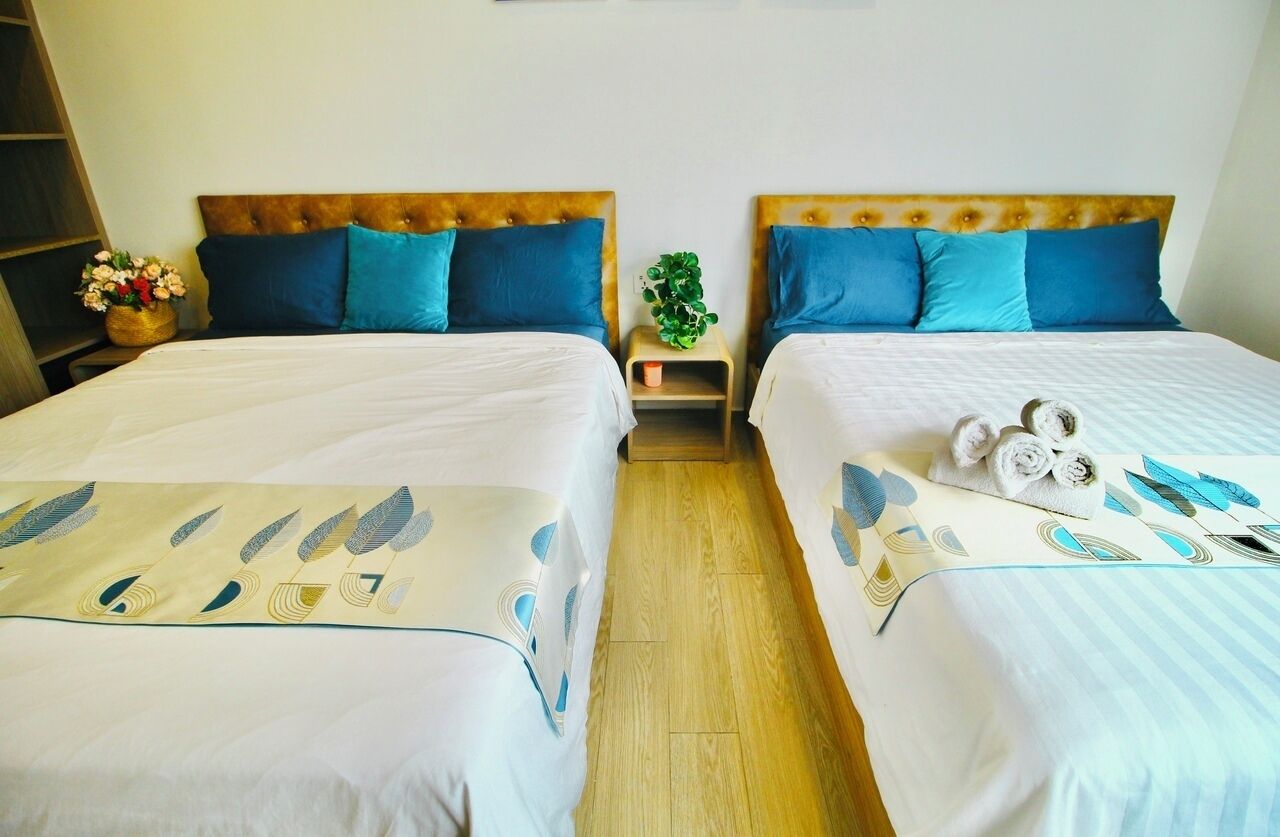 Habitación de hotel con dos camas, cada una con cabecera, almohadas azules y colcha blanca. Hay una planta pequeña sobre una mesita de noche entre las camas, y una cesta de flores a la izquierda.