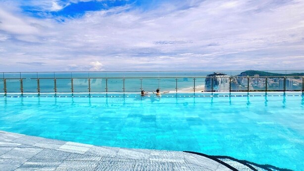 A swimming pool with clear blue water, a glass barrier, and a view of the beach and city skyline. - The Song Vung Tau Apartment (Ho Chi Minh City)