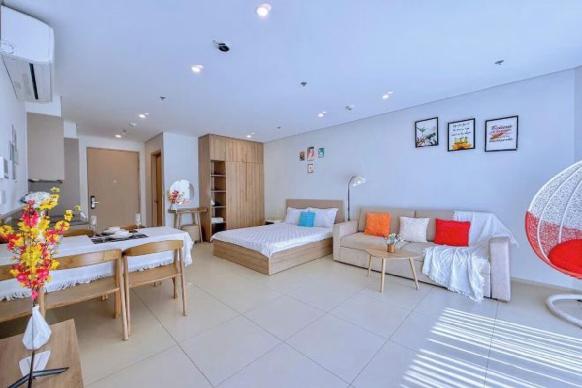 Habitación de hotel moderna con cama, sofá, mesa de comedor y silla. Se observa una mesita con lámpara y planta decorativa.