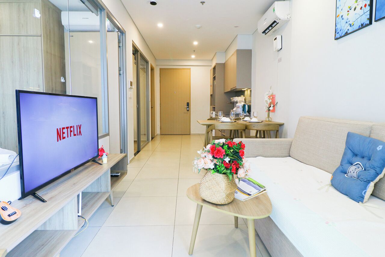 Una sala de estar moderna con un televisor de pantalla plana mostrando 'NETFLIX', un sofá, una mesa de centro con un arreglo floral y un comedor al fondo.