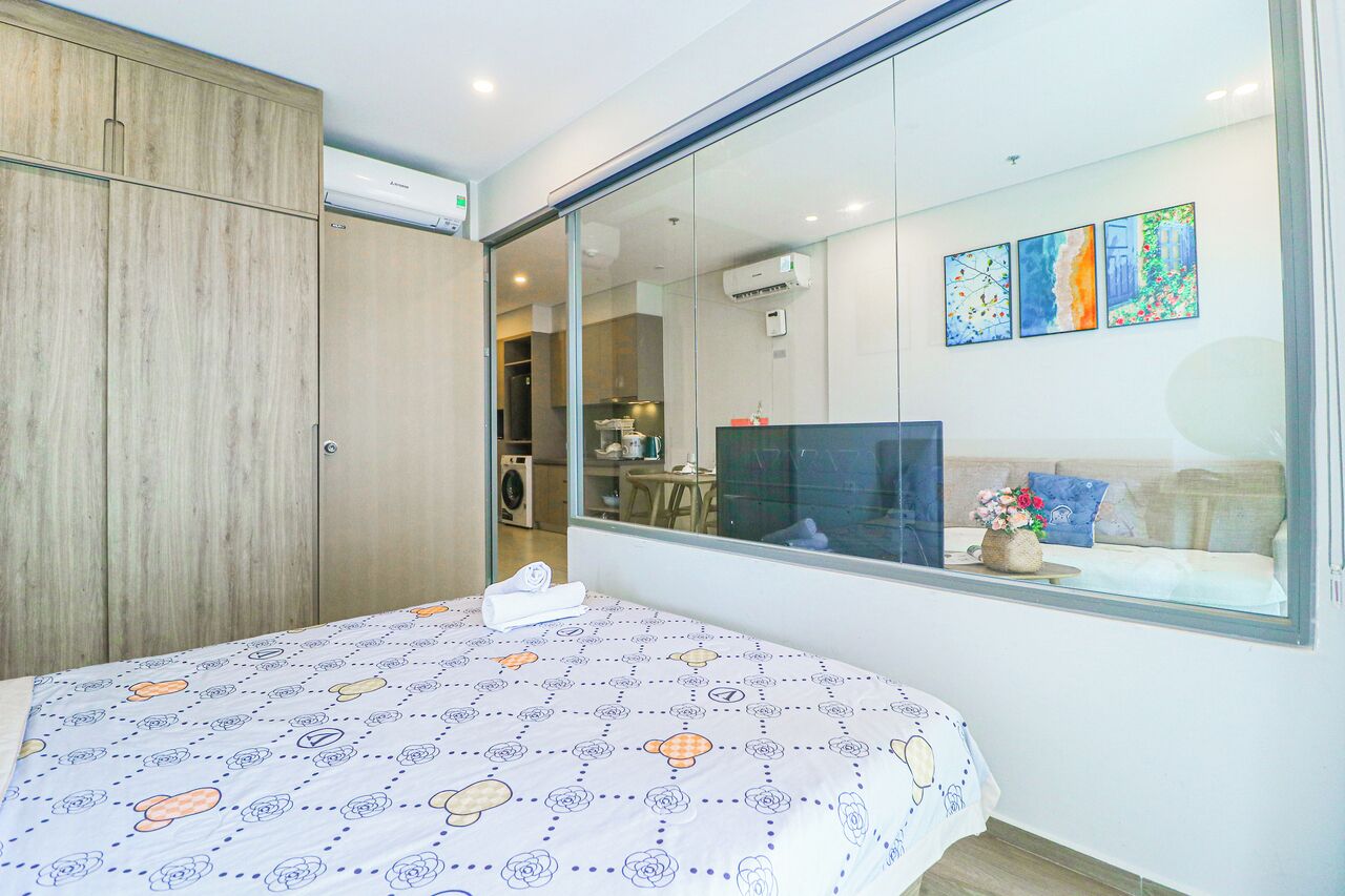 A bedroom with a bed, a wardrobe, a large window, and a view of a kitchen and dining area.
