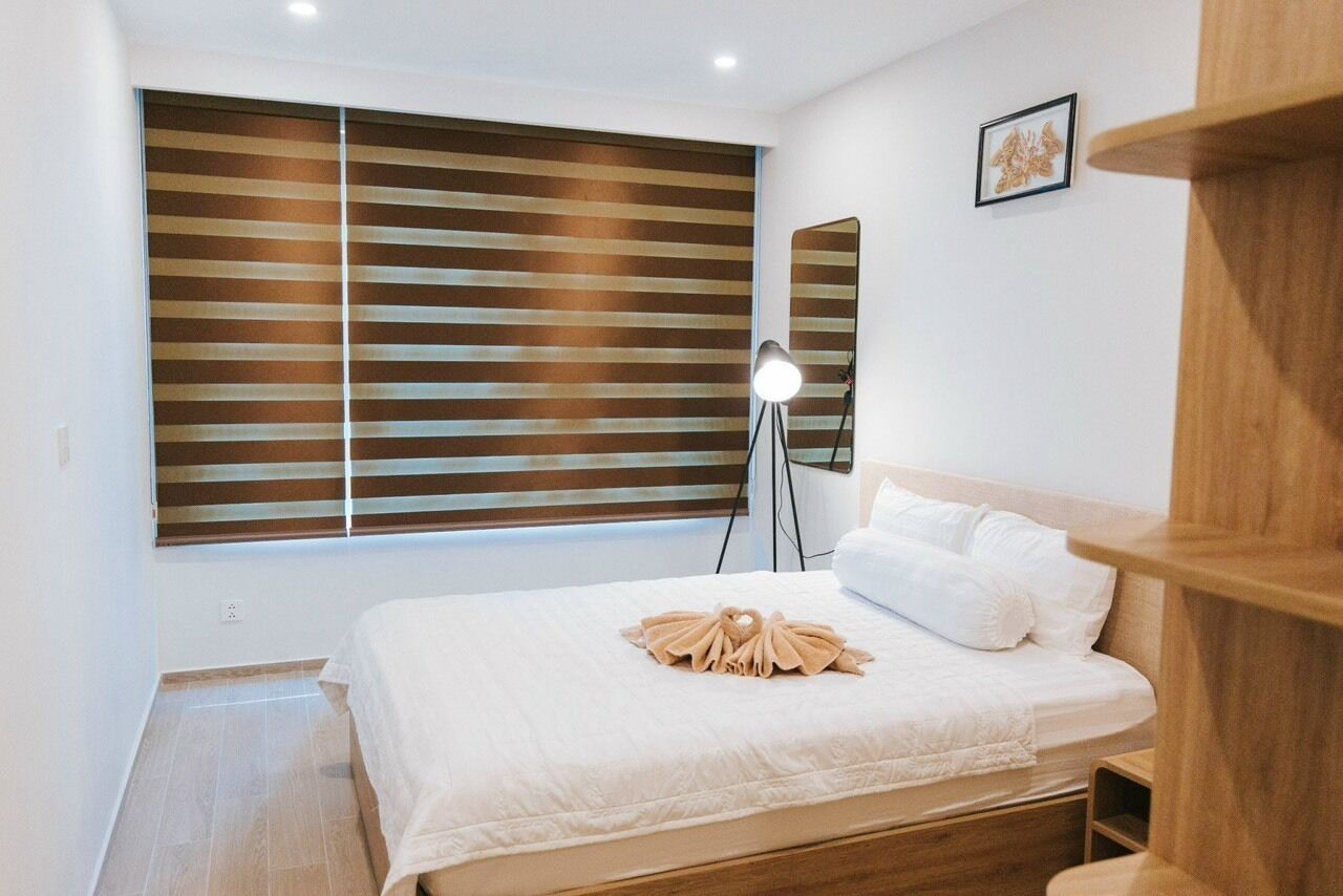 Ein Schlafzimmer mit einem Bett, einer Nachttischlampe, einem Fenster mit Jalousien und einem kleinen Holzregal.