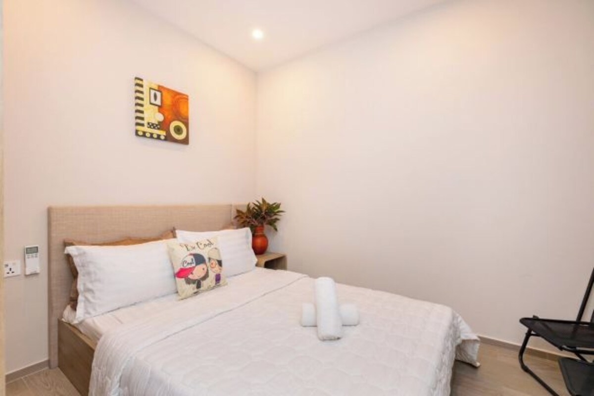 Una cama bien hecha con sábanas blancas, un almohadón con un personaje de dibujos animados, una pequeña planta en maceta y un cuadro colgado en la pared.