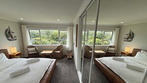 A hotel room with two beds, a small table, two chairs, and large windows offering a view of greenery. - Waiake Beach Apartments (Auckland)