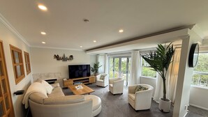 A modern living room with a large L-shaped sectional sofa, a flat-screen TV, and a coffee table. There are two armchairs and a potted plant. - Waiake Beach Apartments (Auckland)