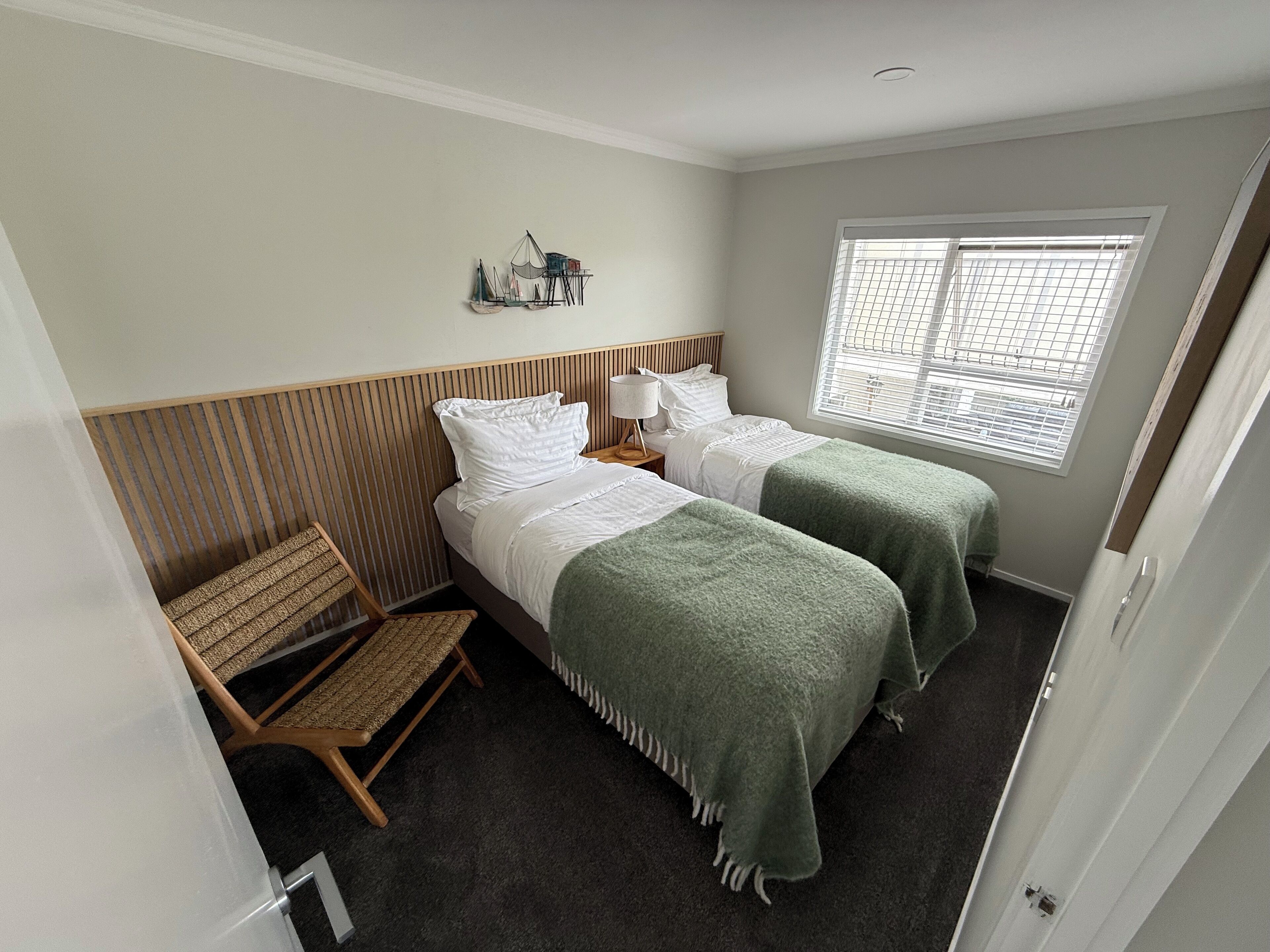 Ein Zimmer mit zwei Betten, einem Fenster mit Jalousien, einem kleinen Tisch mit Lampe und einem Stuhl.