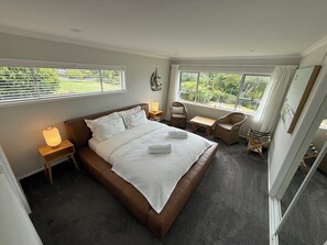 A bedroom with a large bed, two bedside tables, a chair, and a small table. There are windows with curtains and a view of greenery outside. - Waiake Beach Apartments (Auckland)