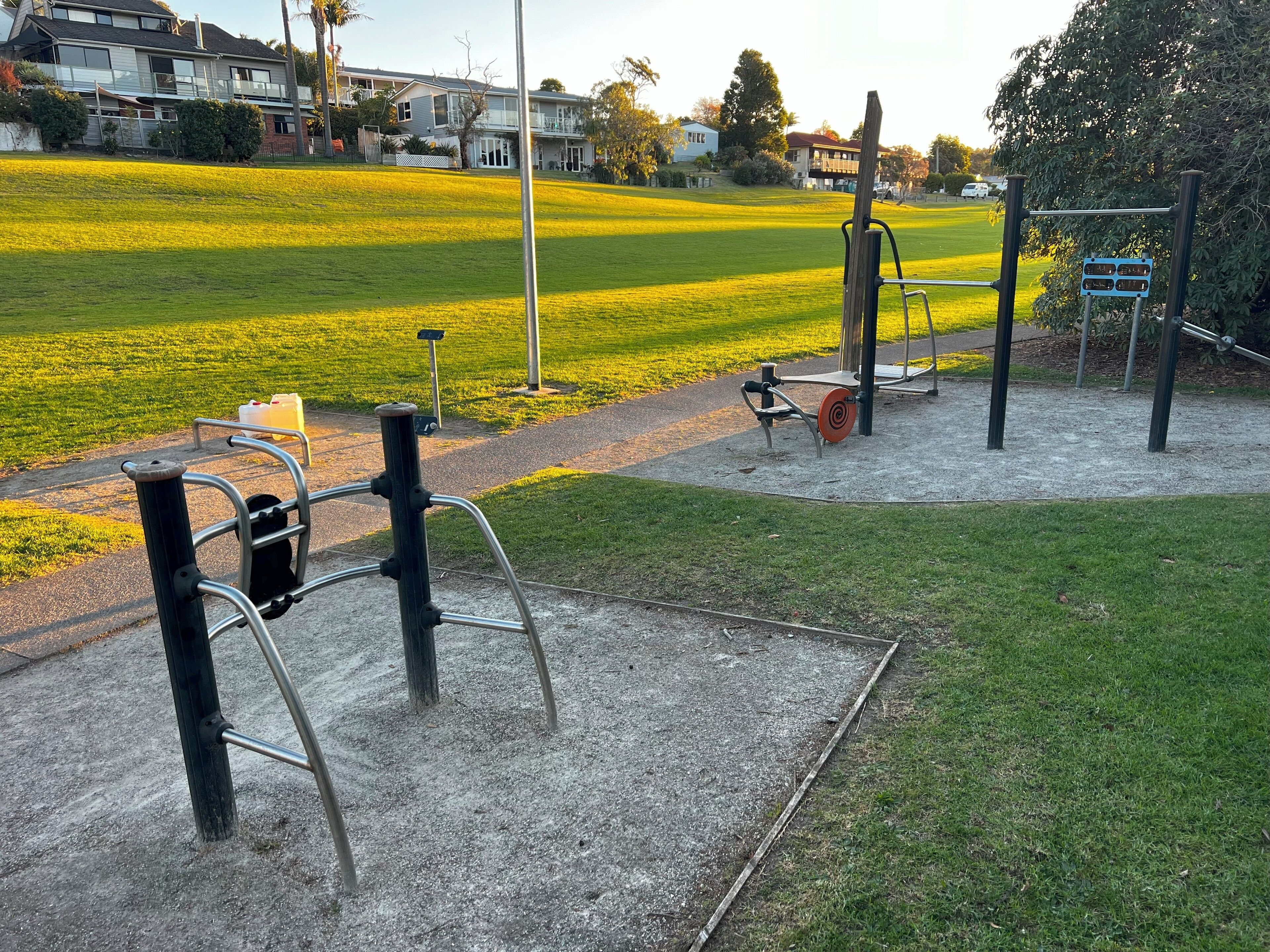 Equipos de ejercicio al aire libre en un parque con un campo de césped y casas al fondo.
