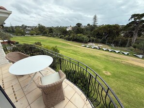 A balcony with wicker furniture and a view of a grassy area with sailboats. - Waiake Beach Apartments (Auckland)