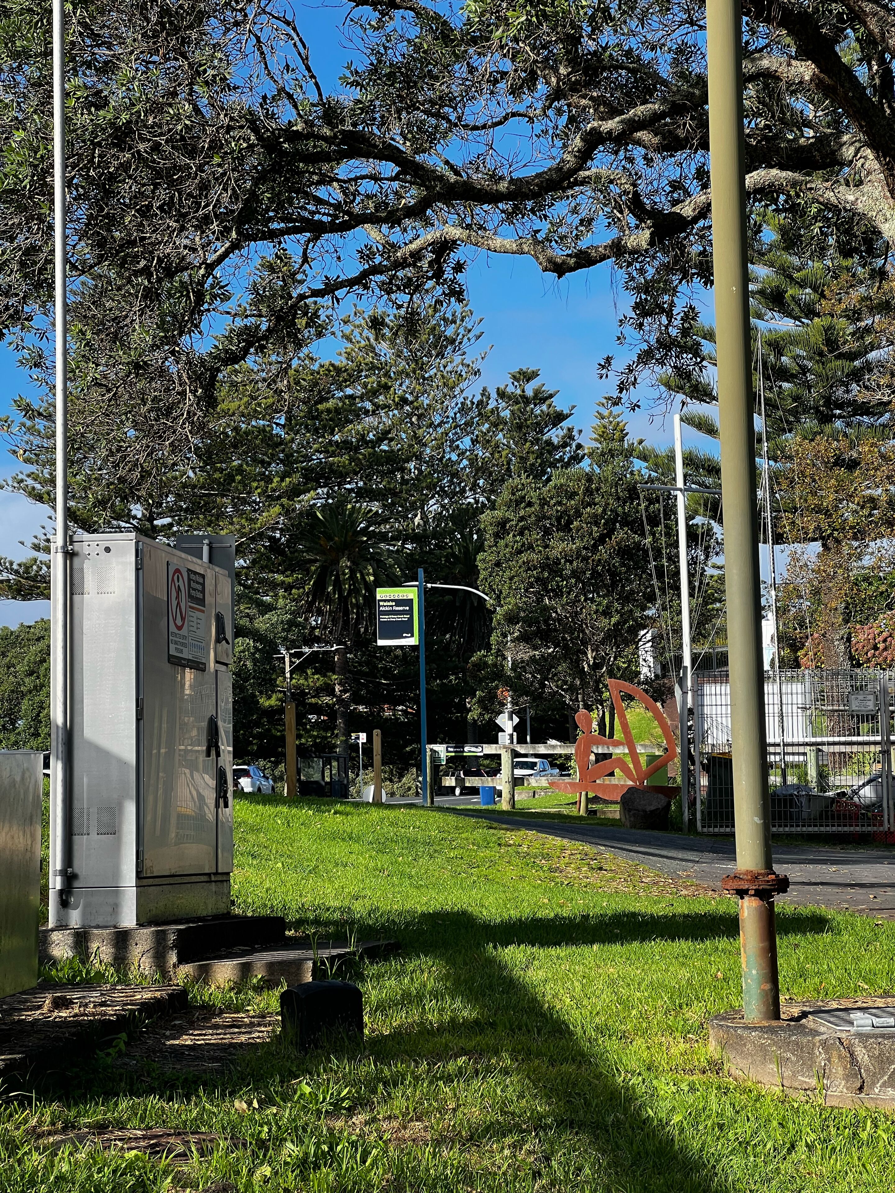 A park with a metal utility box, a sign indicating a walking path, and a metal fence.