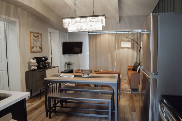 A modern kitchen with a dining area, wooden flooring, a chandelier, a flat-screen TV, and a refrigerator.