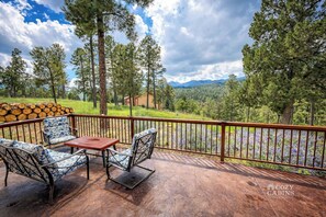 A covered outdoor deck with metal chairs and a table, overlooking a forested area with a mountain in the distance.