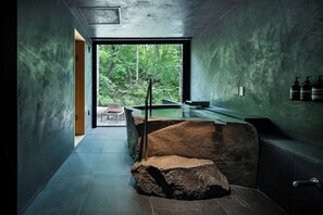 Un baño moderno con una gran escultura de roca, un banco y vistas a un bosque a través de un ventanal.
