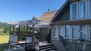 Une terrasse extérieure couverte, aménagée avec des meubles en bois, une pergola et offrant une vue sur une maison dotée de grandes fenêtres.