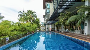 A swimming pool with a tiled floor, surrounded by greenery and a building with a modern design. - Luxury Everything Walking Distance c112 (Rawai)