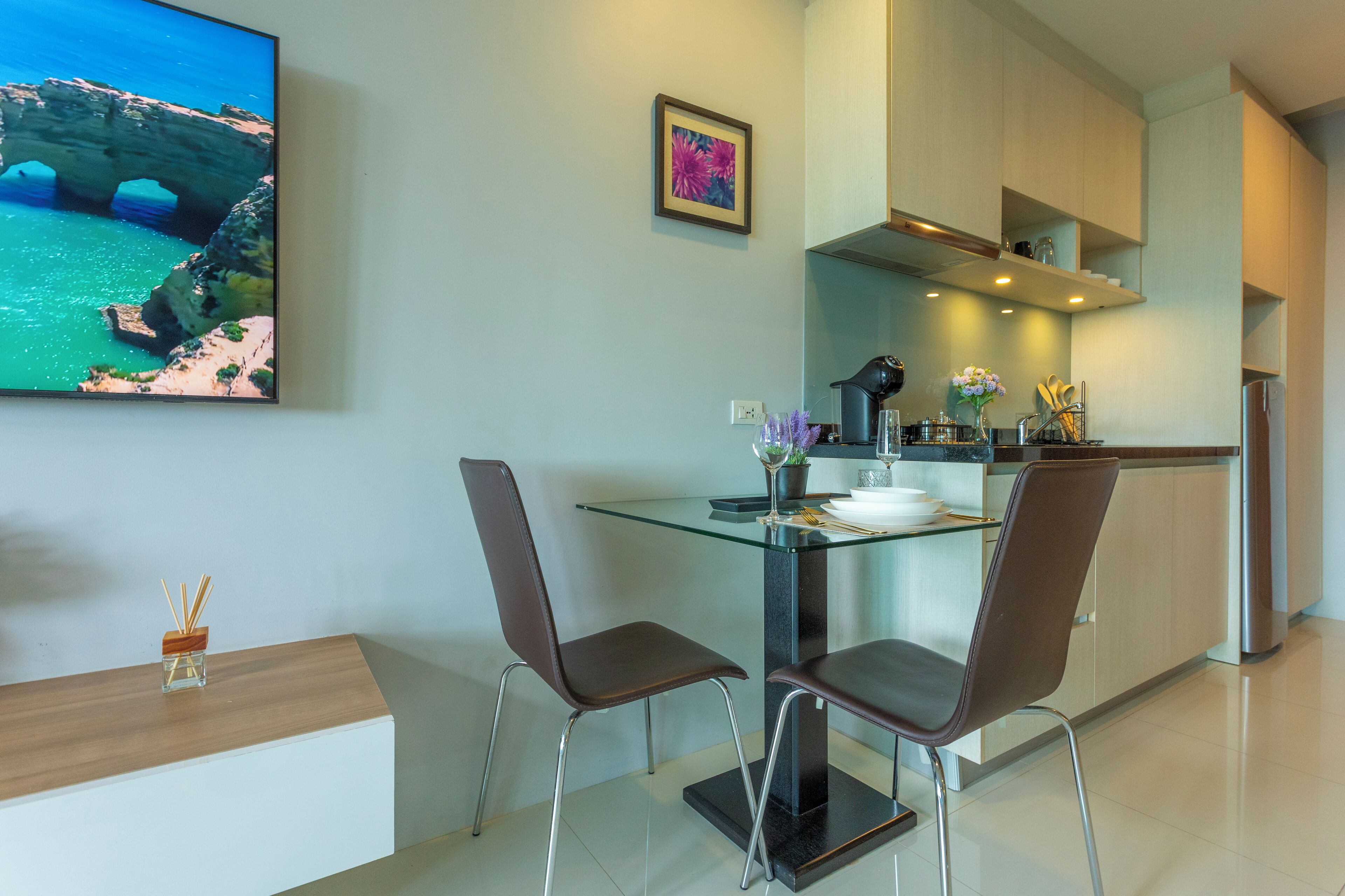 A modern kitchen with a glass dining table, two chairs, and a large flat-screen TV mounted on the wall.
