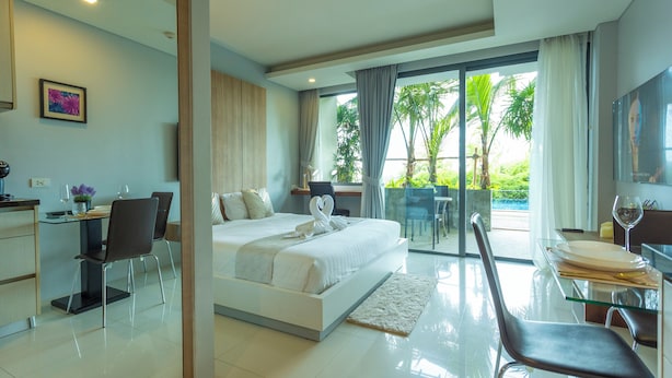 A modern hotel room with a large bed, a desk with a chair, a dining area with a table and chairs, and a balcony with a view of greenery. - Luxury Everything Walking Distance c112 (Rawai)