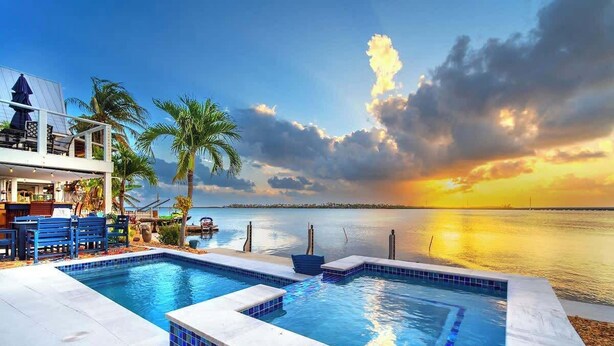 A swimming pool with a view of the sunset over water, a deck area with chairs, and a building with a balcony.