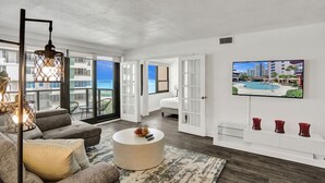 A modern living room with a large window, a round coffee table, and a flat-screen TV on the wall.