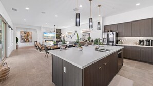 Una cocina moderna con una isla grande, muebles oscuros, electrodomésticos de acero inoxidable, iluminación colgante y un comedor con sillas y una mesa.