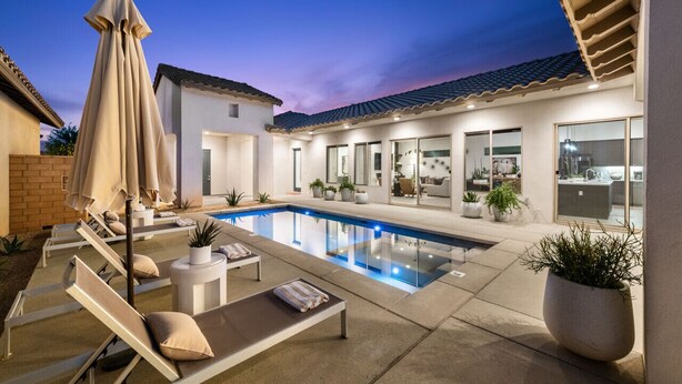 Un área de piscina moderna con sillones de descanso y sombrillas, una piscina cristalina y una casa con ventanas amplias y techo de tejas.