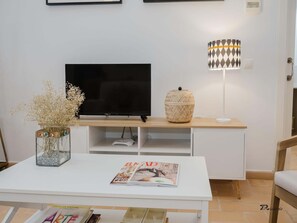 Un salon avec un téléviseur à écran plat posé sur un meuble TV en bois, une table basse blanche avec des magazines, un vase décoratif contenant des fleurs séchées et un lampadaire à motifs.