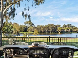 A dining table with chairs, a bowl, and glasses, overlooking a lake with trees and a grassy area.