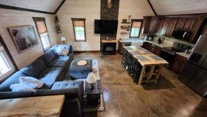 Ein gemütliches Wohnzimmer mit Sofa, Hocker und Couchtisch, einem Kamin und einer Küchenzeile mit Holzschränken und Geräten.