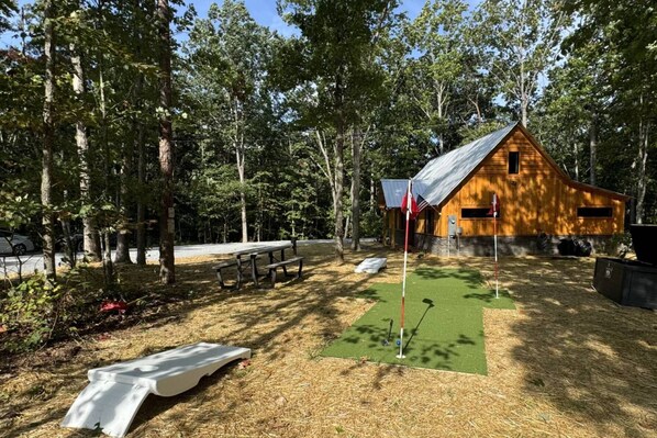 Una cabaña rústica con un felpudo verde y un asta de bandera, rodeada de árboles y mesas de picnic.