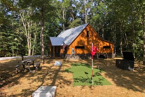 Una cabaña rústica con techo de metal, rodeada de árboles. Hay mesas de picnic y una esterilla verde en el suelo.