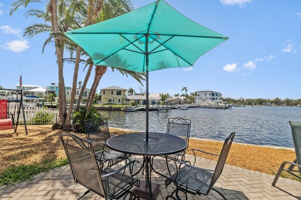 A round metal table with four chairs and a green umbrella, set on a paved area with a view of a lake and buildings in the background. - Waterfront with Pool, Ping Pong table, 3 Kayaks, Fishing Pier, Private Access to the Gulf (Port Richey)