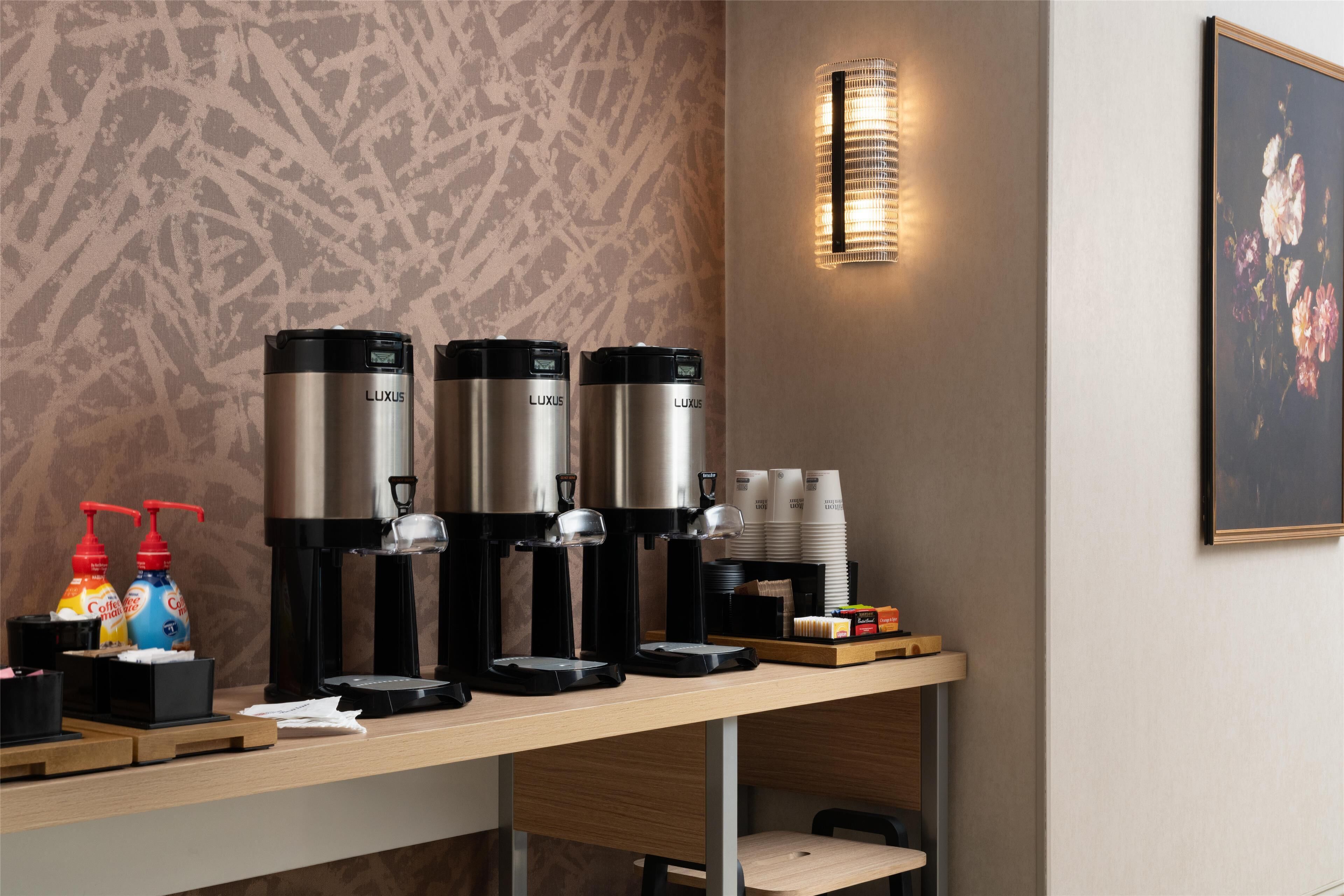 Angolo caffè con tre macchine da caffè LUXUS, una lampada a parete e un quadro floreale incorniciato.