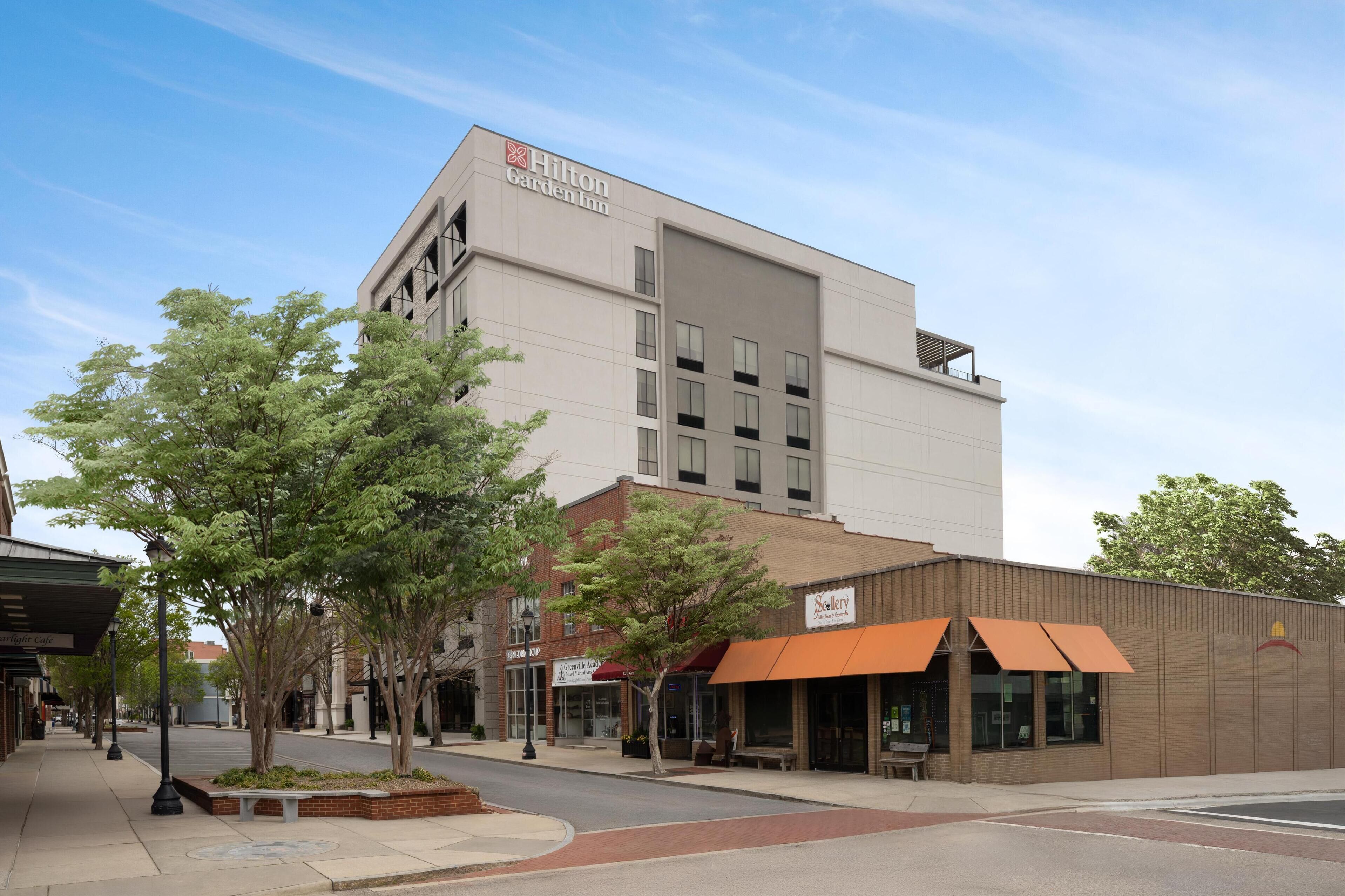 Ein Hilton Garden Inn Hotel mit moderner Fassade, einem Baum-bewachsenen Bürgersteig und Geschäften, darunter eine Galerie und ein Restaurant.