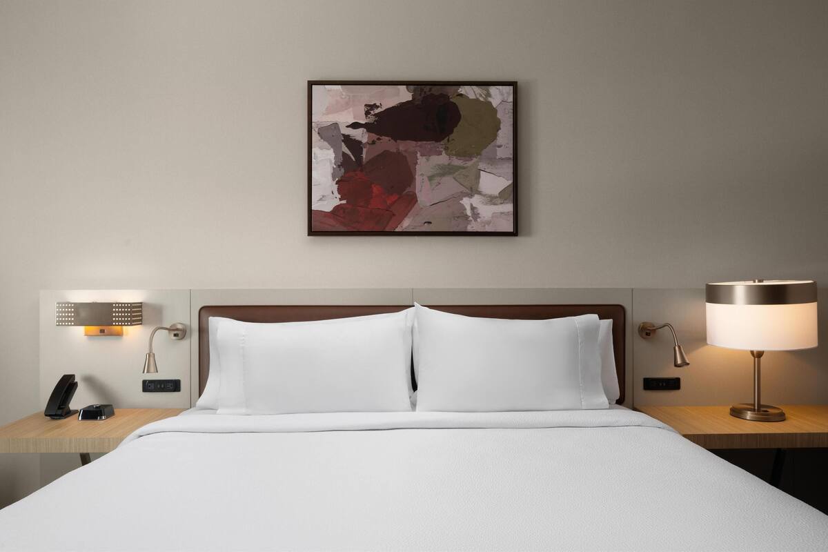 Une chambre d’hôtel avec un lit soigneusement fait, une table de chevet avec une lampe, un téléphone et un tableau abstrait accroché au mur.