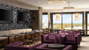 A modern dining area with cushioned seating, tables, and a large window offering a view of a tower and landscape. - Hilton Garden INN Greenville University (Greenville)
