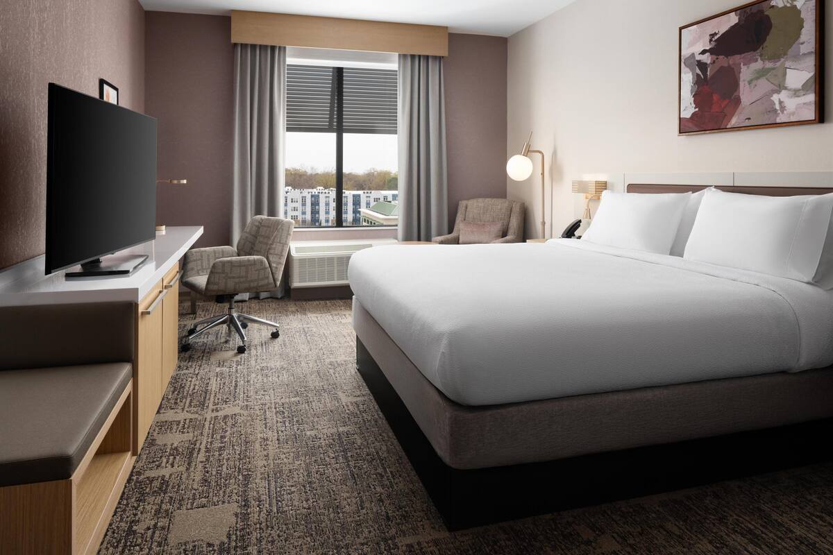 Une chambre d’hôtel avec un grand lit, un bureau doté d’une télévision à écran plat, une chaise et une fenêtre donnant sur la ville.