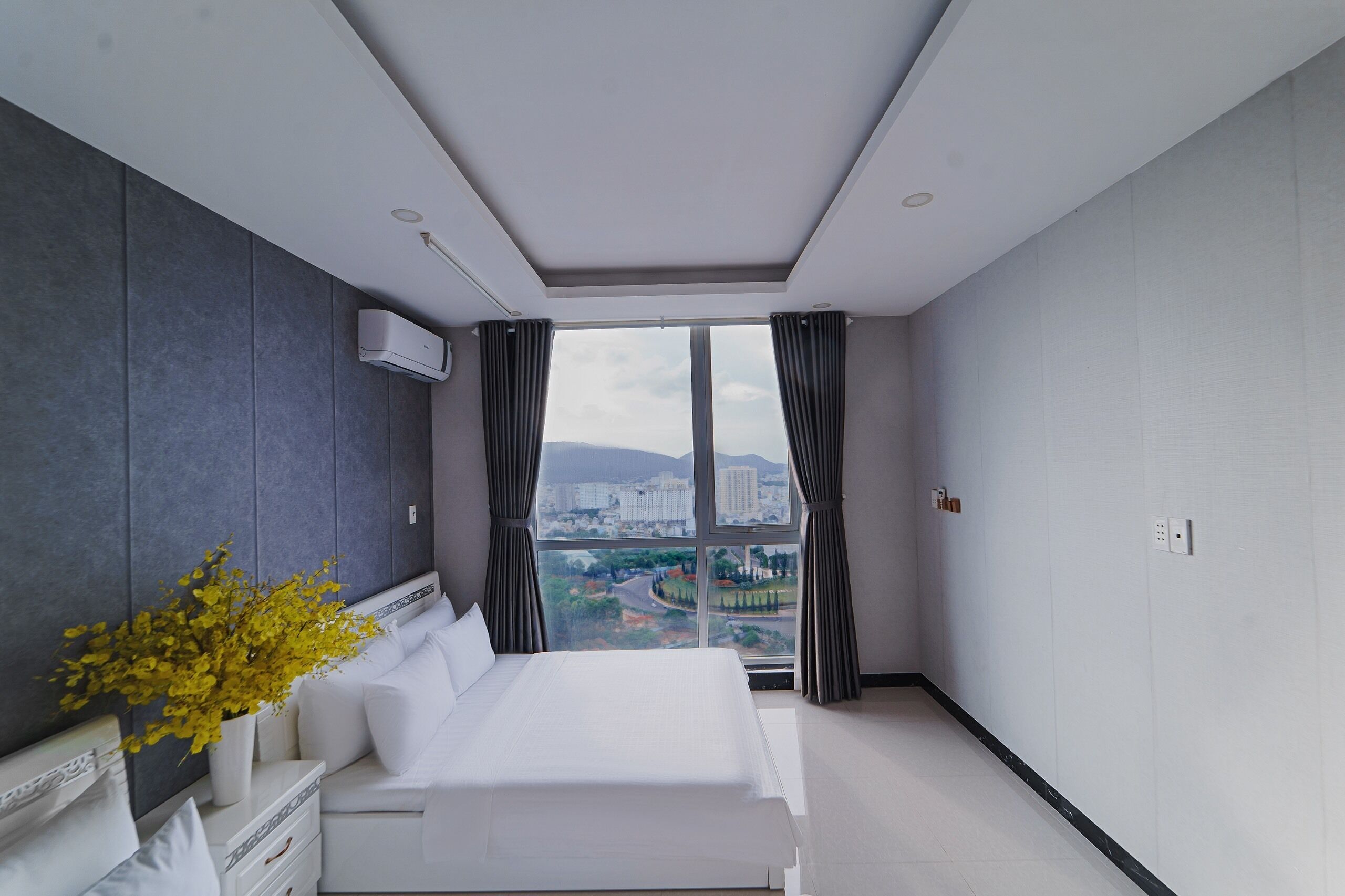 Ett modernt hotellrum med en stor säng, ett nattduksbord, en krukväxt och utsikt över en stadssiluett genom stora fönster.