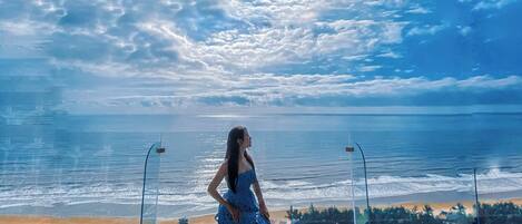 A woman in a blue dress standing on a balcony overlooking a beach with a clear sky.
