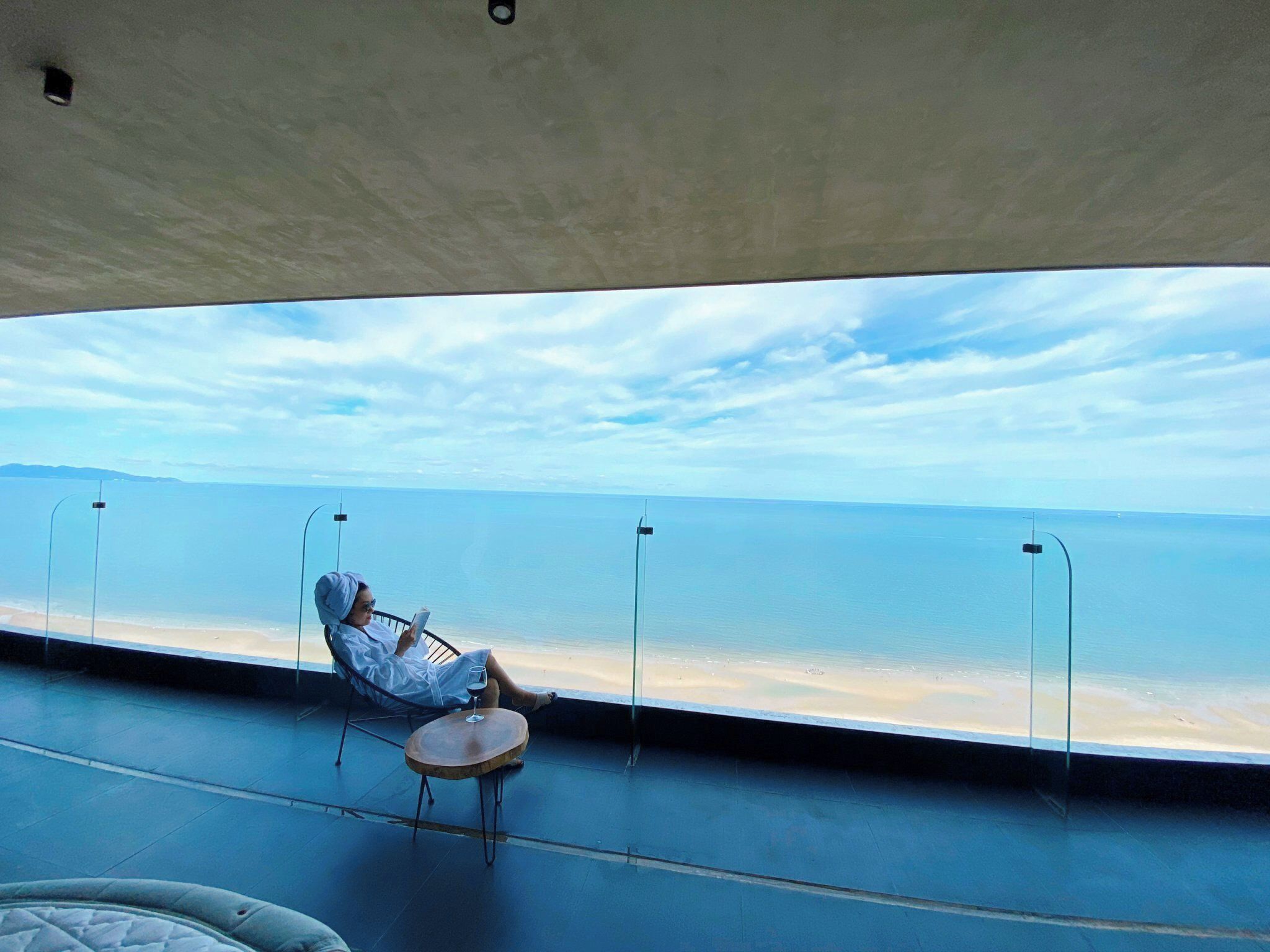 A person seated on a chair by a large window overlooking a beach.