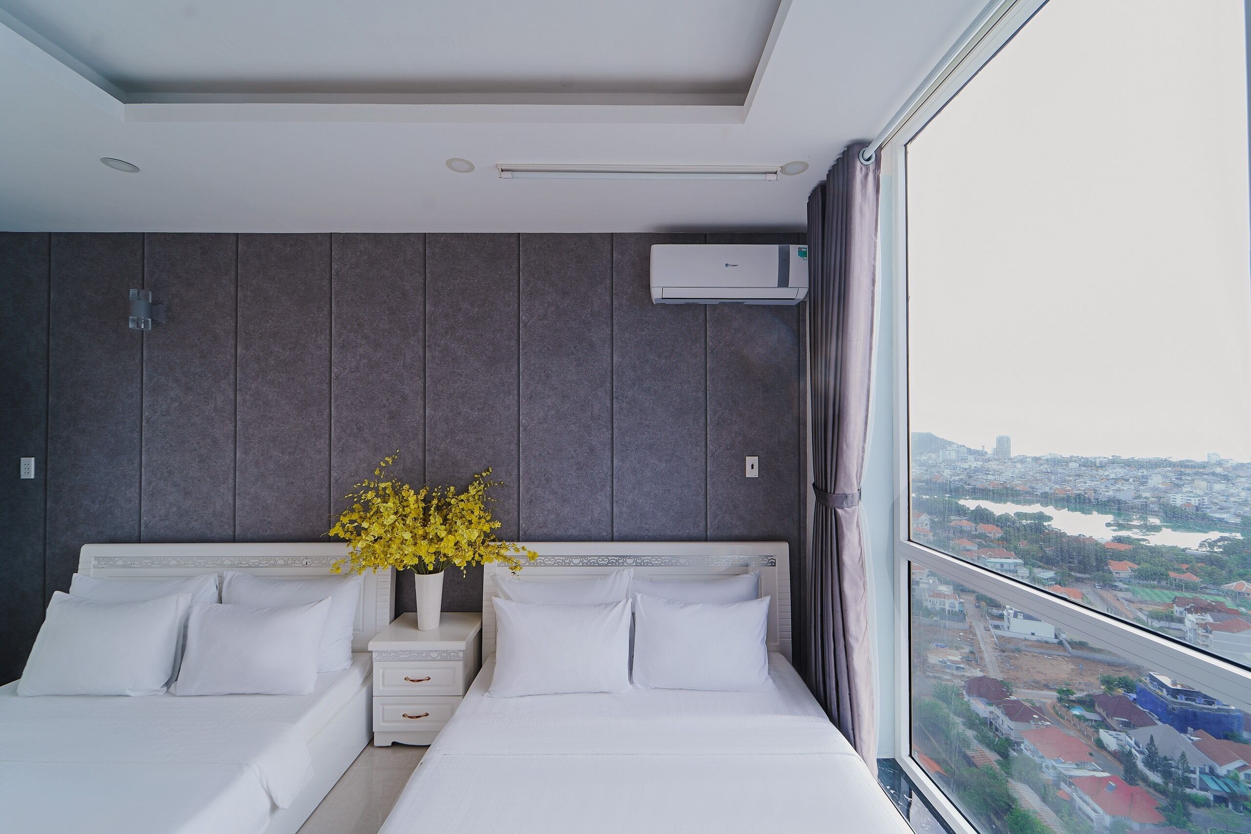Une chambre d’hôtel moderne avec un grand lit, une table de chevet, un vase rempli de fleurs jaunes et une vue sur le paysage urbain, visible à travers une grande fenêtre.