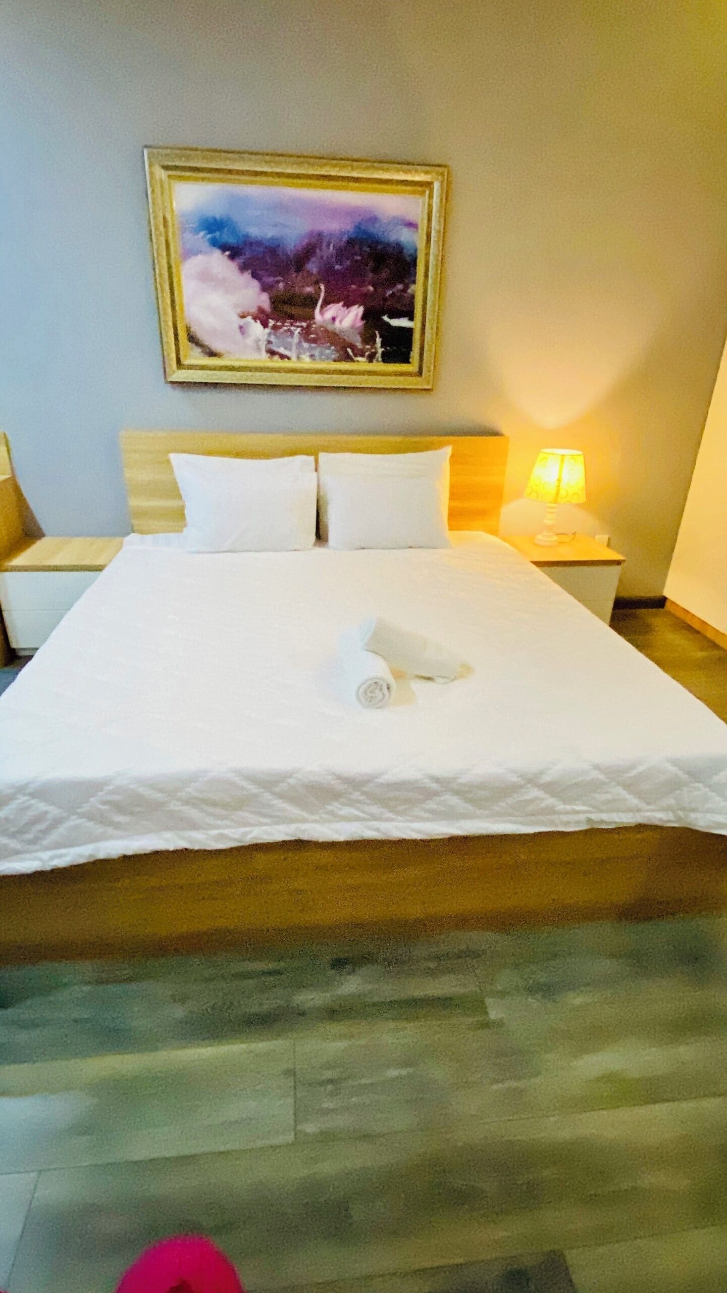 Un lit bien fait, avec du linge de lit blanc et une serviette pliée, une table de chevet avec une lampe et un tableau encadré au-dessus du lit.