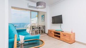 A modern living room with a blue sofa, a glass coffee table, a wooden TV stand, and a large window offering a view of the sea. - WELCS APARTAMENTO 181 EMP con vistas al mar (Rosas / Roses)