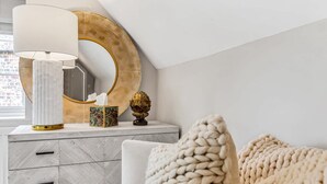 A modern bedroom with a white bed, a grey dresser with drawers, a large mirror with a gold frame, a white lamp, and a decorative vase.
