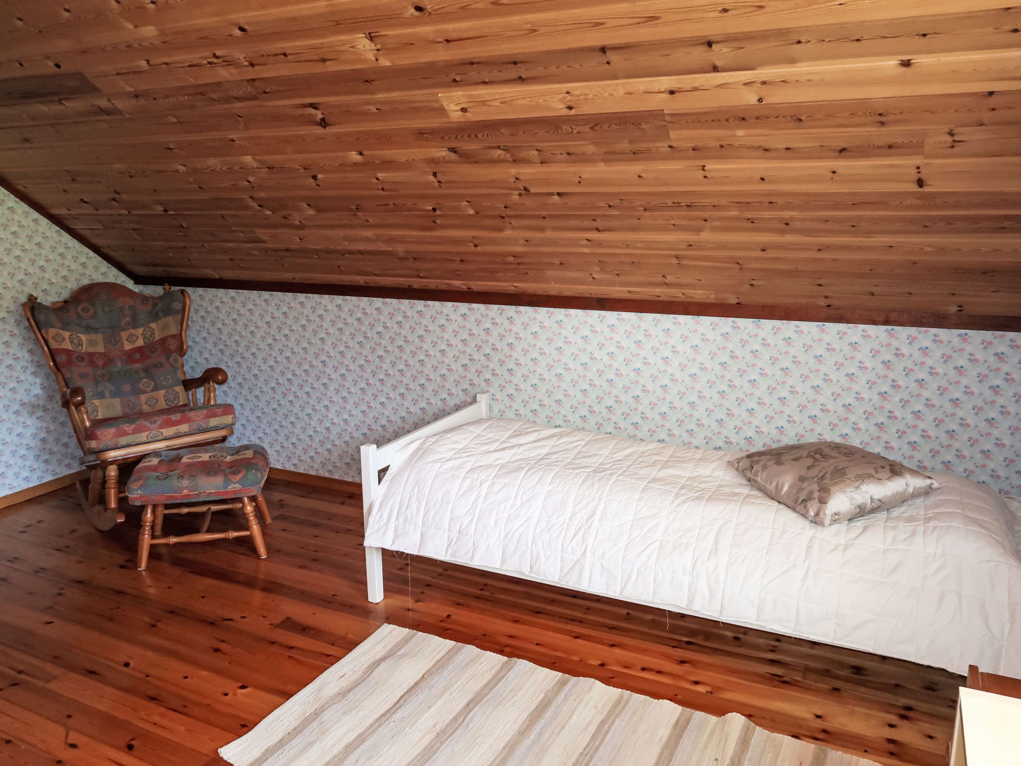 Een slaapkamer met een houten plafond, een enkel bed met een witte quilt, een gestreept fauteuil en bloemenbehang.